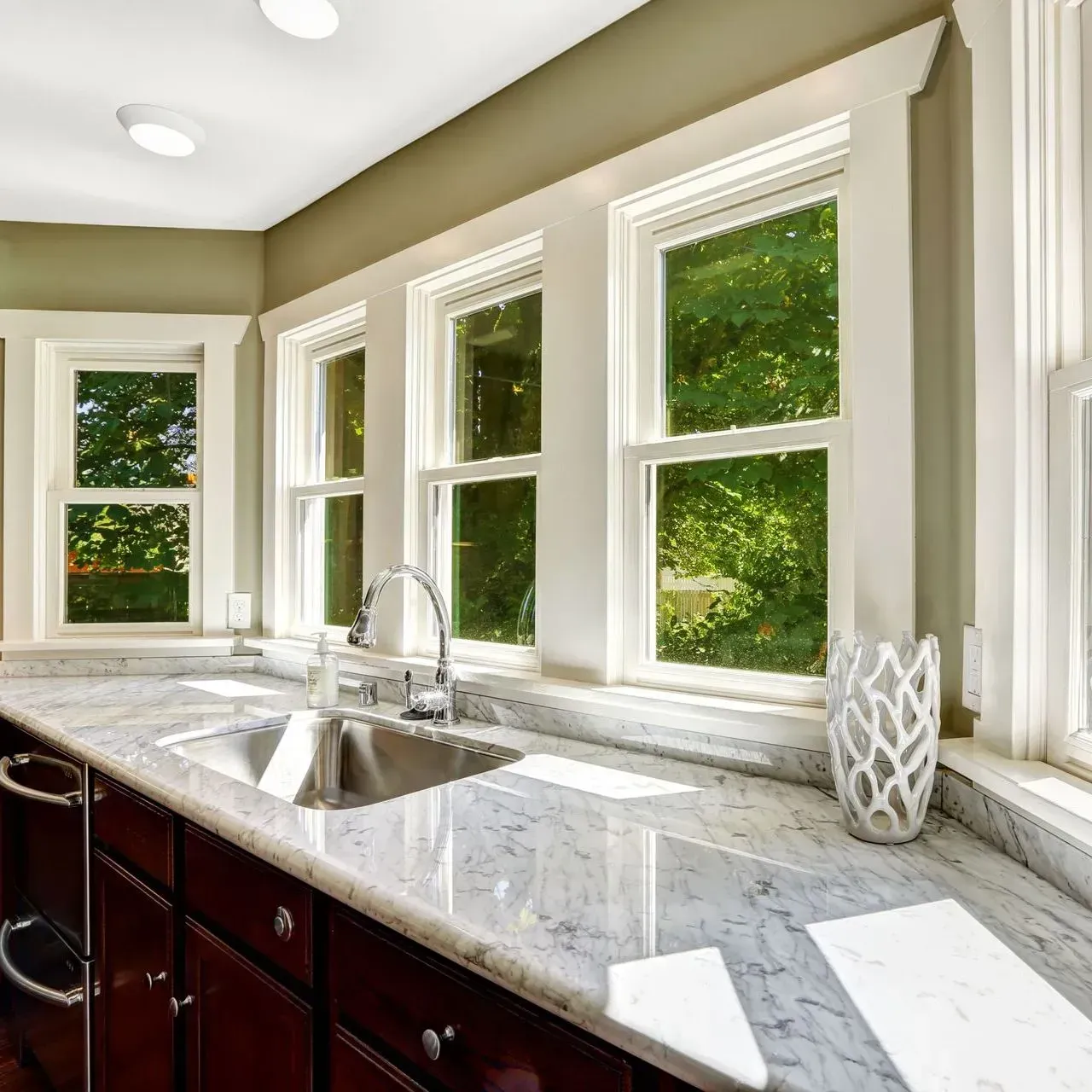 A modern kitchen with marble countertops, a stainless steel sink, and large windows with white trim overlooking trees.