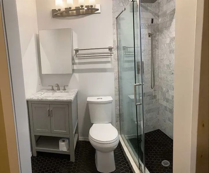 A modern bathroom with a grey vanity, white toilet, and a glass-enclosed shower with dark tile floors and marble walls.