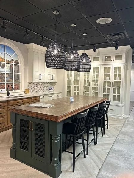 A kitchen showroom featuring a large dark green island with a wood top, black spindle stools, and three woven pendants.