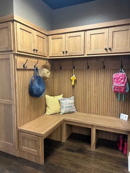 An L-shaped wooden mudroom bench with upper cabinets, coat hooks, and two pillows, set against a paneled wall.