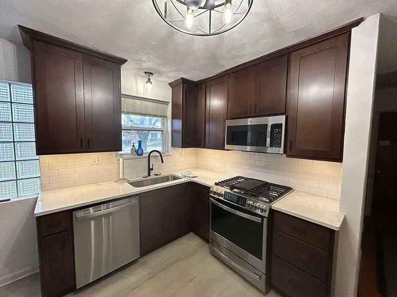 A modern kitchen with dark brown cabinets, light countertops, a stainless steel oven, dishwasher, and glass block window.