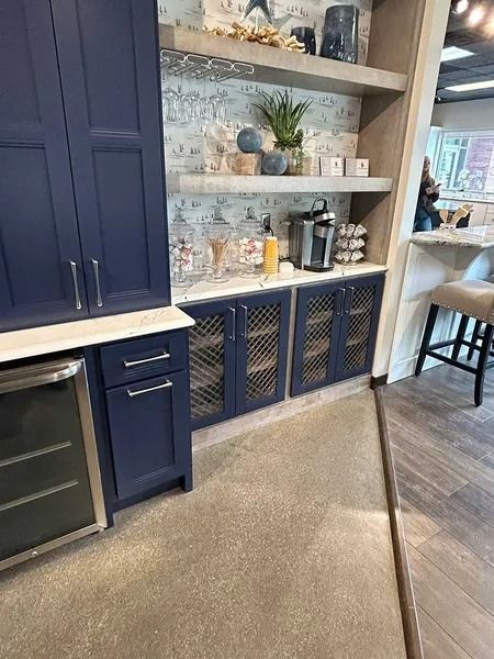 Navy kitchen cabinetry with glass-paneled doors, shelves, and a coffee station, situated next to a bar area with stools.