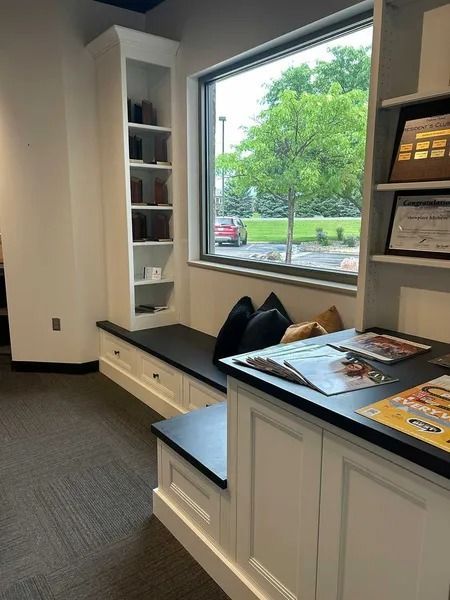 White built-in cabinetry with a window seat and shelving, featuring black countertops and cushions in a bright office area.