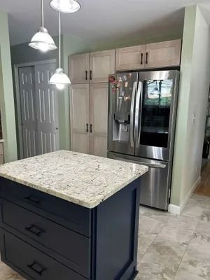 A kitchen island with dark blue cabinets and speckled granite countertops sits before light wood cabinets and a fridge.