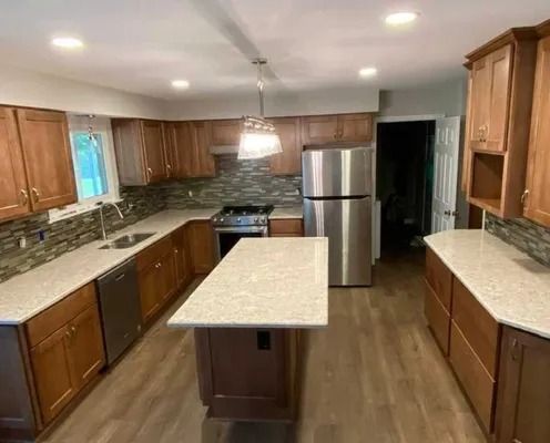 A modern kitchen with wood cabinets, light quartz countertops, a central island, stainless steel appliances, and wood floors.