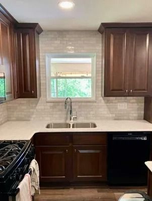 A kitchen with dark wood cabinets, white countertops, a sink under a window, and a tiled backsplash.