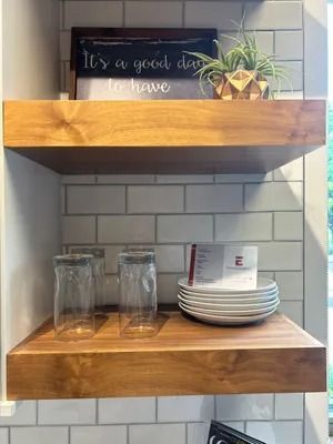 Two floating wooden shelves against a white subway tile wall holding a sign, a small plant, two glasses, and white plates.