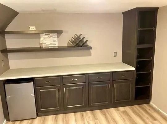 A home bar area with dark cabinetry, open shelving, a white countertop, and an under-counter refrigerator on light floors.