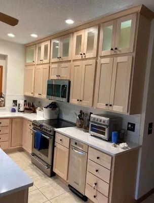 A modern kitchen with light wood cabinets, glass-front upper storage, stainless steel appliances, and white countertops.