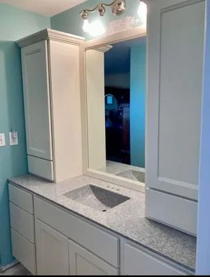 A bathroom vanity with light cream cabinets, a speckled grey countertop, a rectangular mirror, and a three-bulb light fixture.