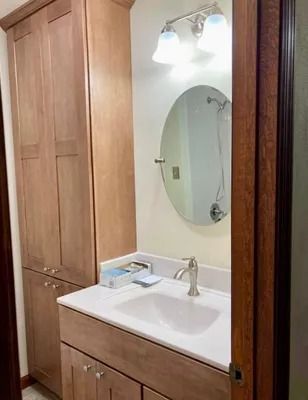 A wooden bathroom vanity with a white countertop, oval mirror, and wall light, viewed through a doorway.