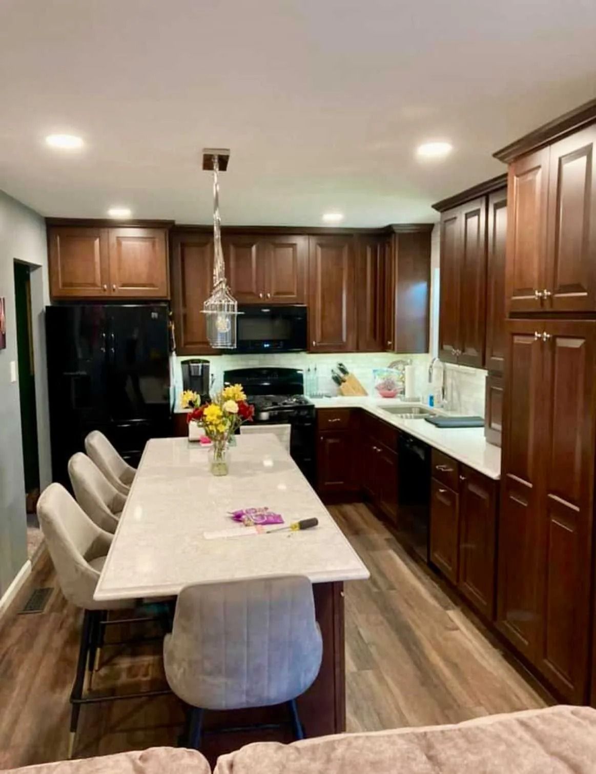 A kitchen with dark wood cabinets, a long white island with four gray stools, black appliances, and light wood flooring.