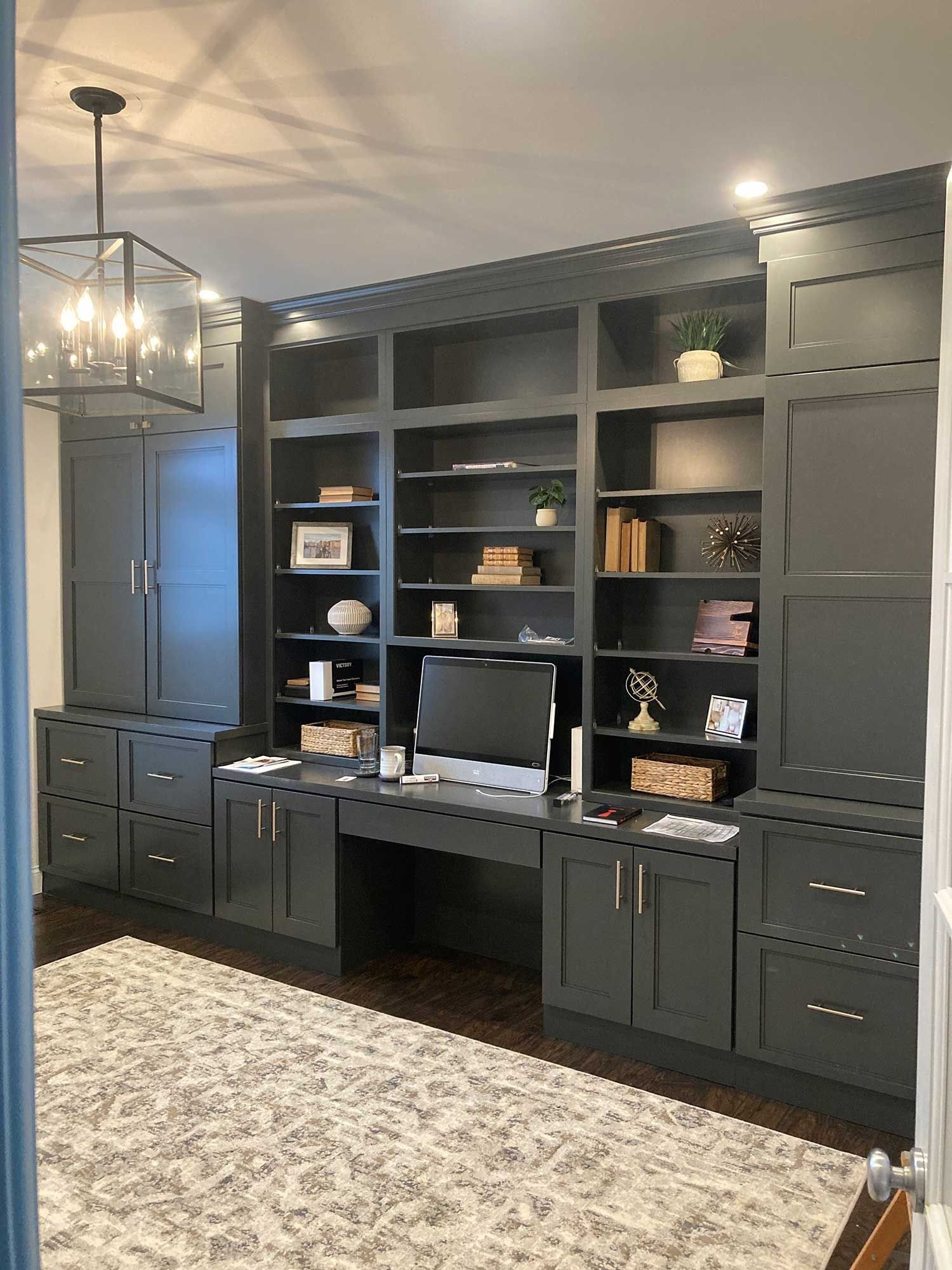 A home office with floor-to-ceiling charcoal grey built-in cabinetry, shelving, a desk with a computer, and a patterned rug.