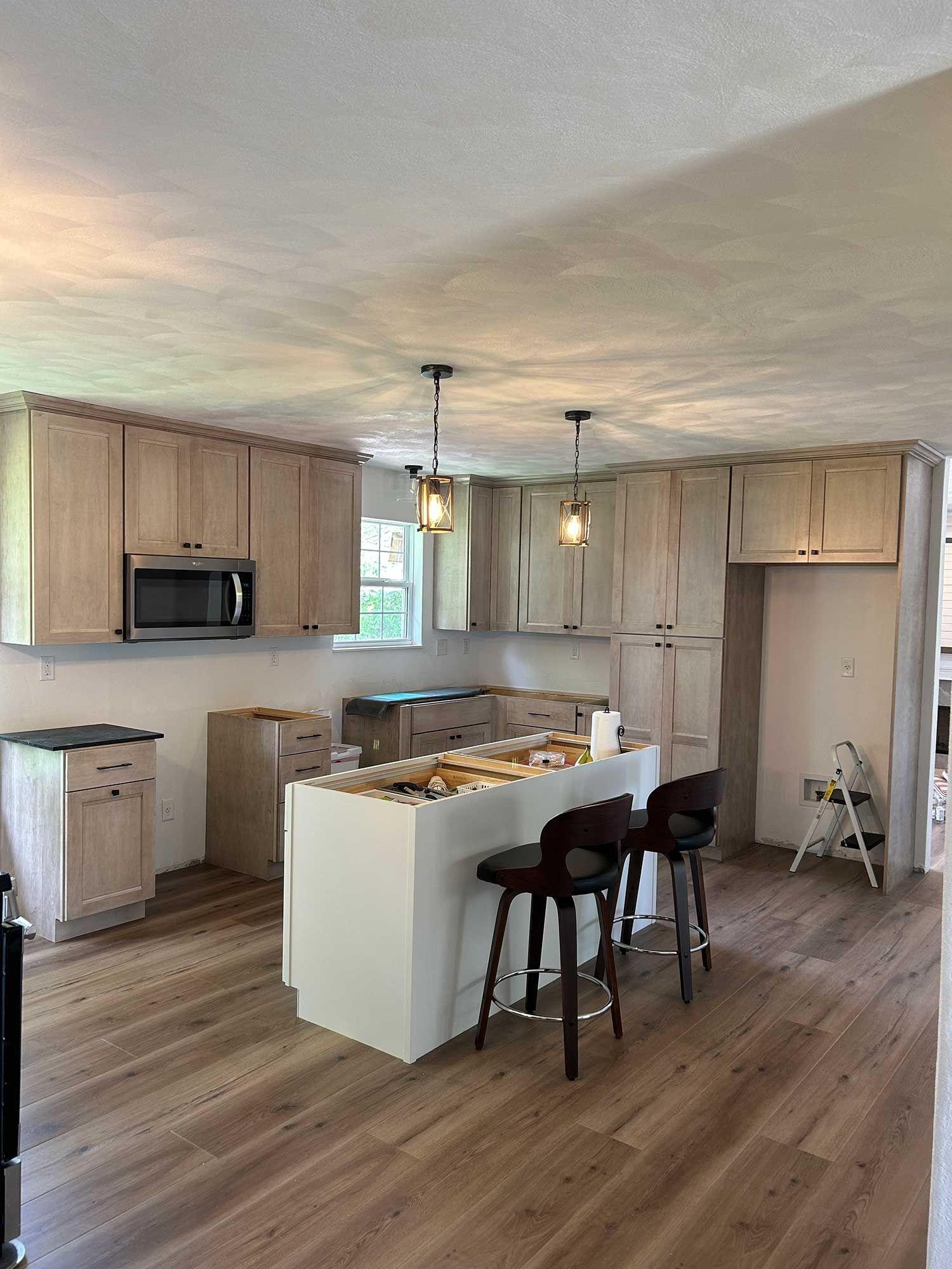 A kitchen under renovation with light wood cabinets, a white center island with two stools, and wood-look laminate flooring.