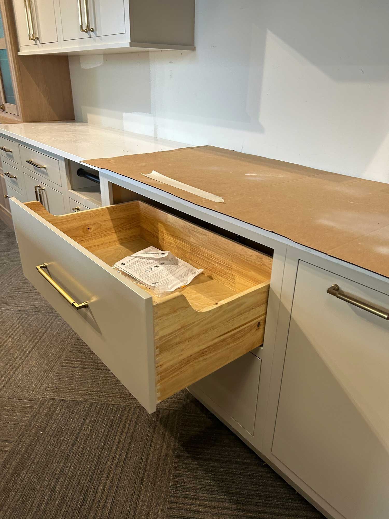 An open wooden drawer with a gold handle on a light-colored kitchen cabinet base in a showroom setting.