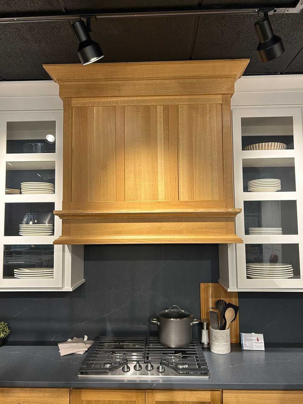A wooden range hood sits between white cabinets above a stovetop with a pot against a dark grey backsplash.