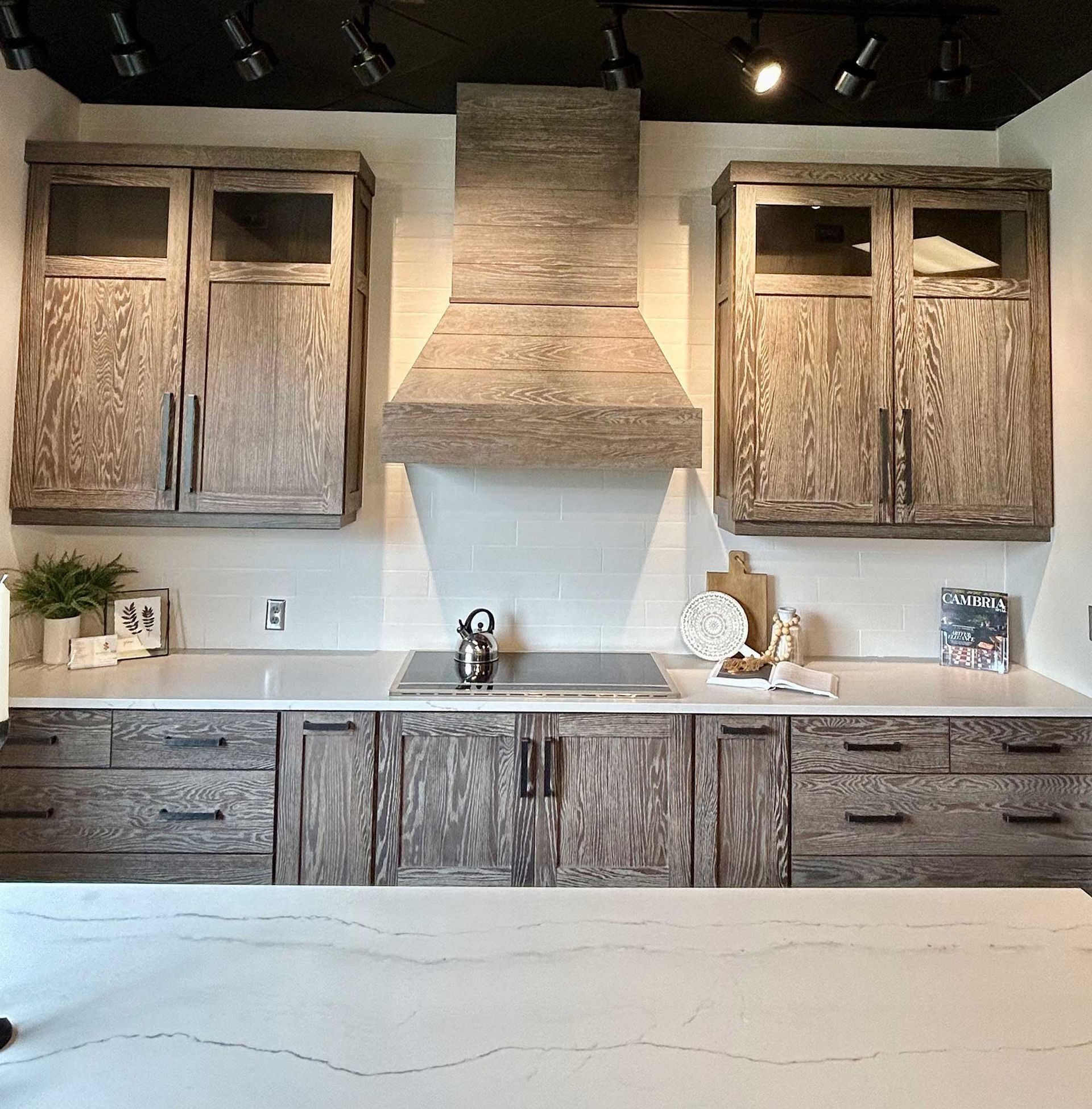 A kitchen featuring rustic wood cabinets, a matching vent hood, white countertops, and a black ceiling with track lights.