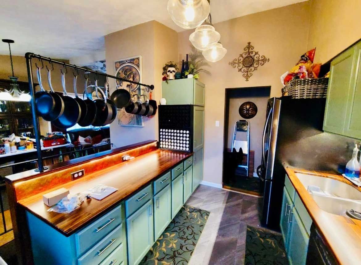 A kitchen with light green cabinets, a wood countertop, a hanging pan rack, and a dark refrigerator next to a doorway.
