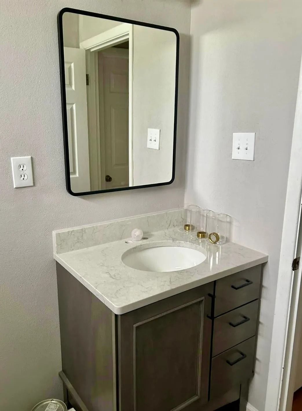 A bathroom vanity with a gray cabinet, white marble-style countertop, circular sink, and a black-framed rectangular mirror.