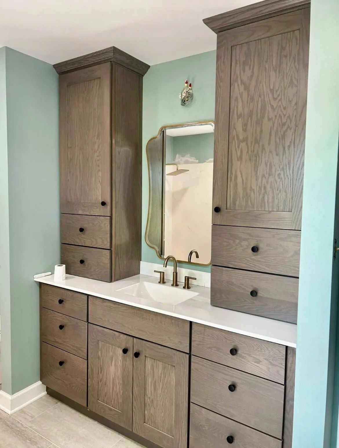 A bathroom vanity with a white countertop, wooden cabinets, and a decorative mirror against light green walls.