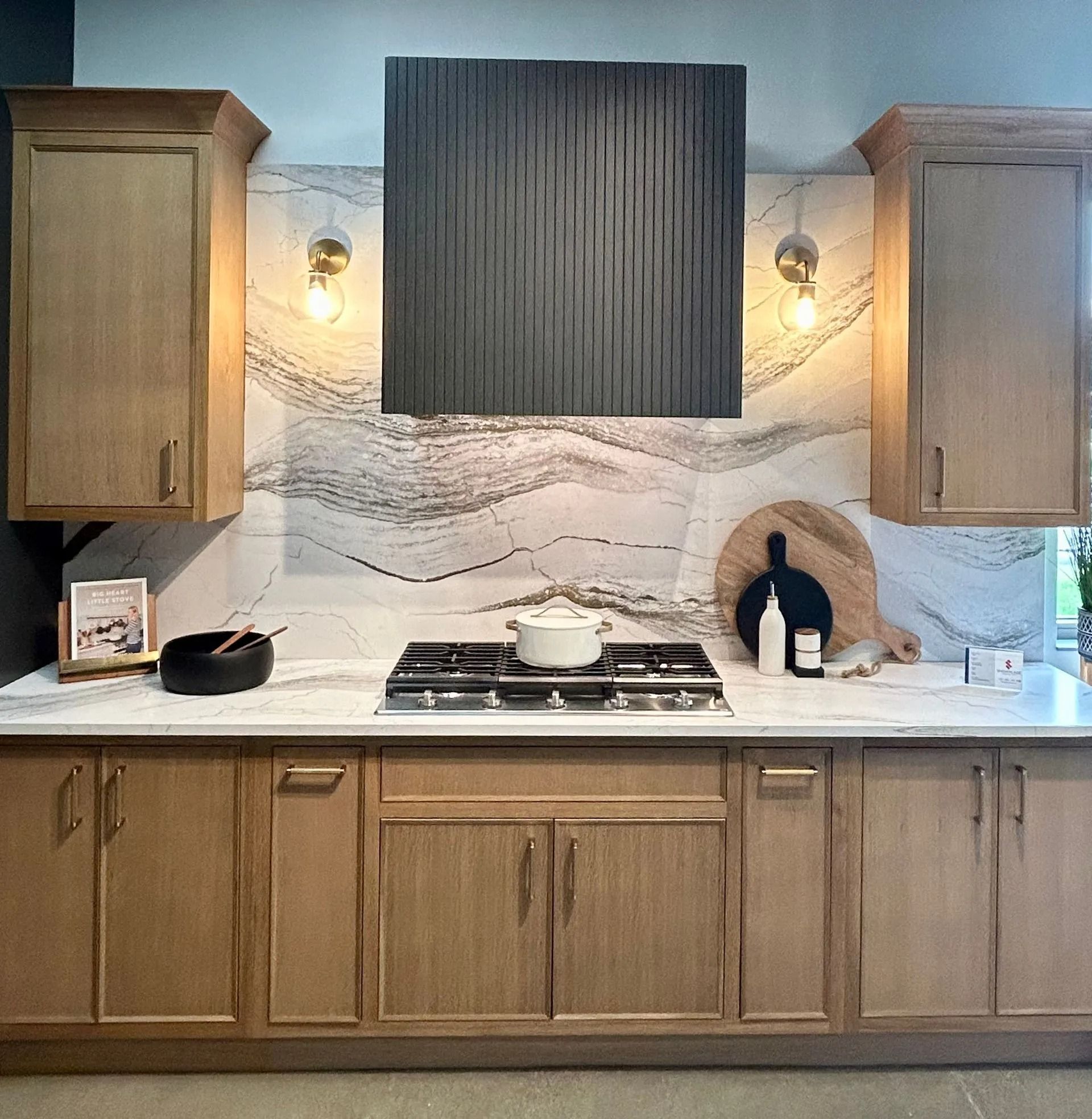 A modern kitchen with wood cabinets, white stone countertops, a large marble backsplash, and a dark, textured range hood.