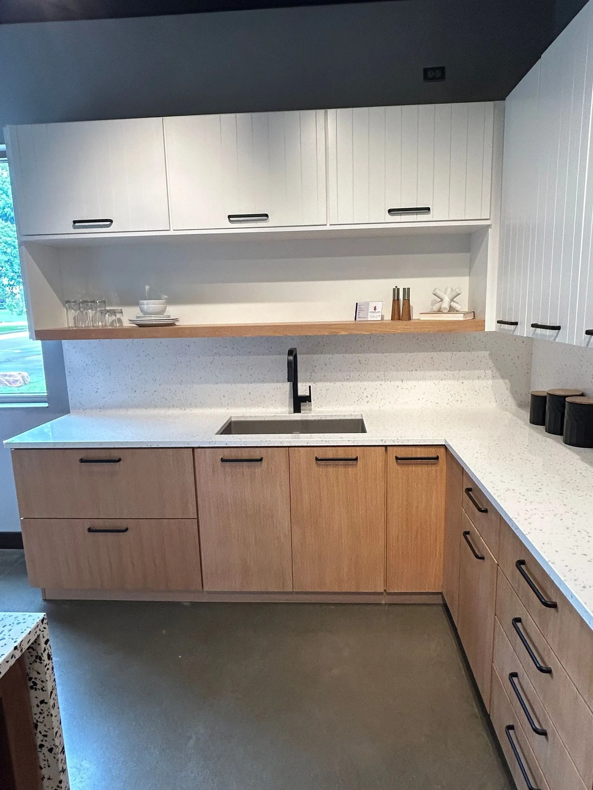 A modern kitchen with light wood cabinets, white speckled countertops, a dark faucet, and white upper cabinets.