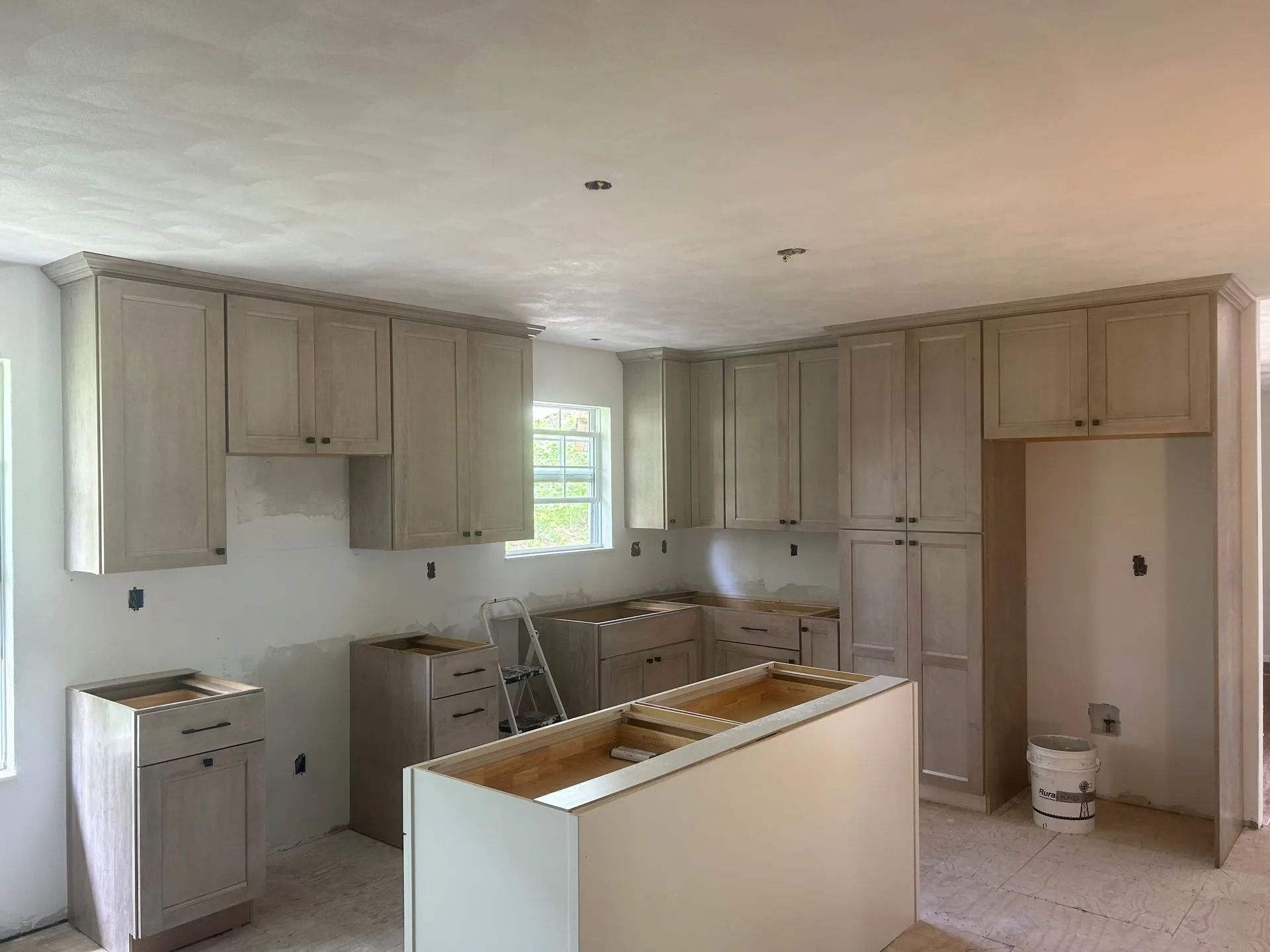A kitchen under construction with light wood cabinets, unfinished flooring, and an open central island base.