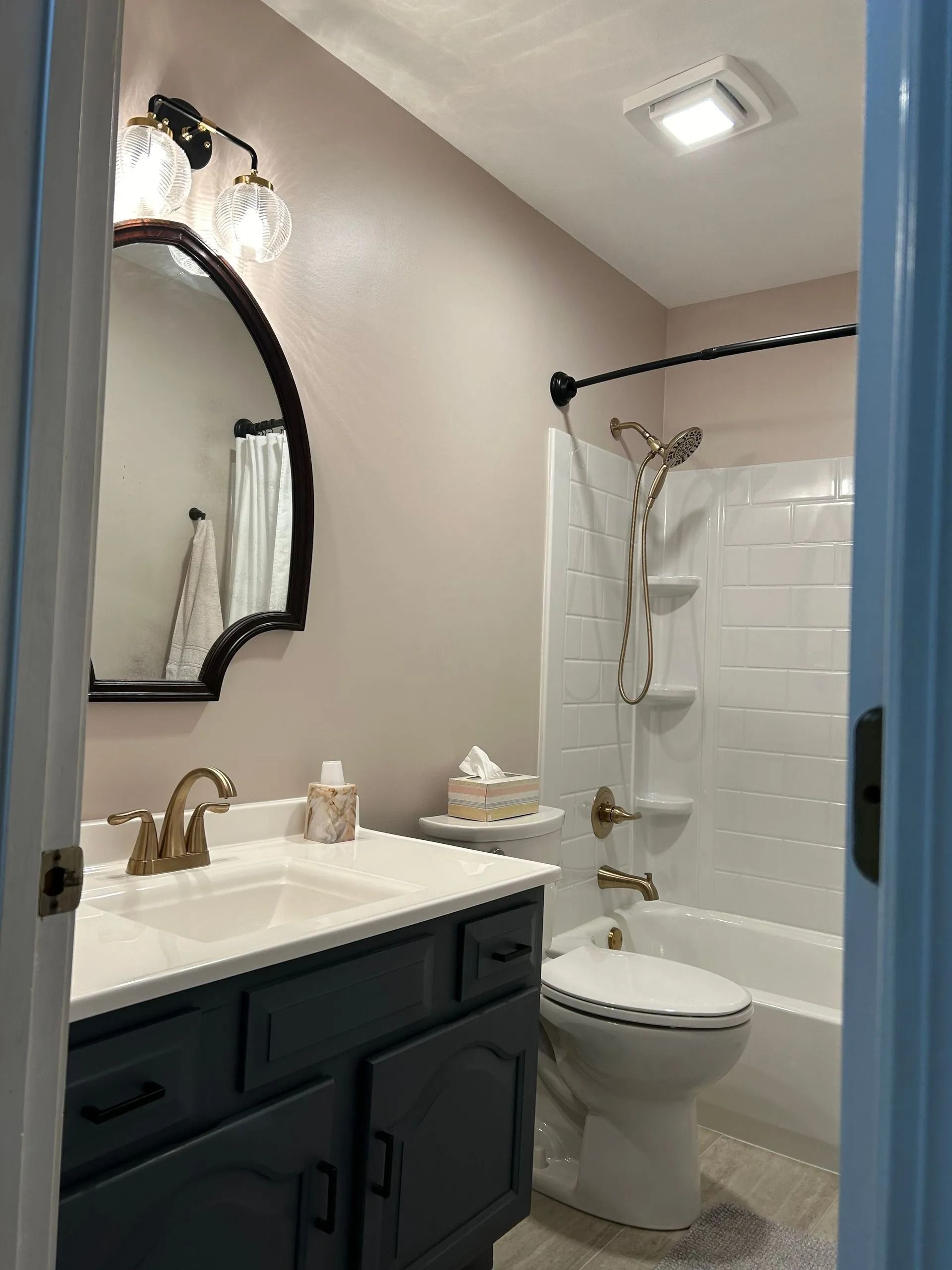 A bathroom with a dark gray vanity, arched mirror, neutral walls, and a white shower-tub combination with gold fixtures.