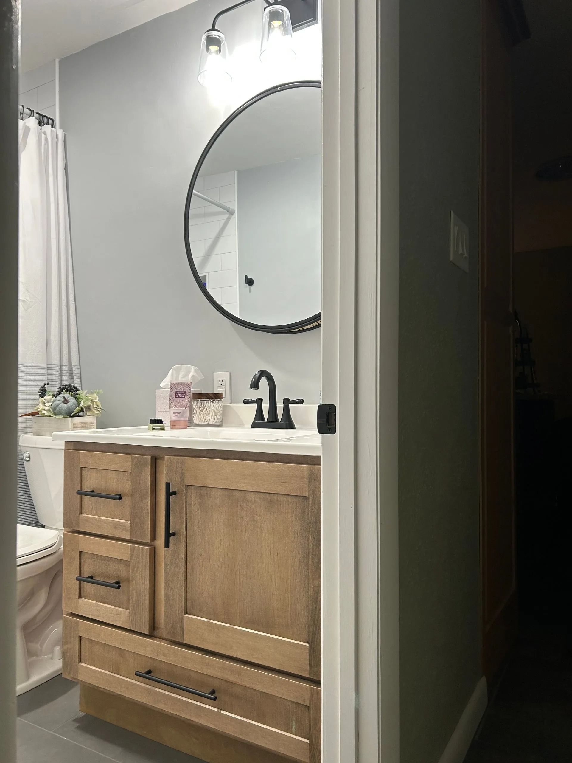 A bathroom vanity with a light wood cabinet, matte black hardware, and a round mirror against a gray wall.