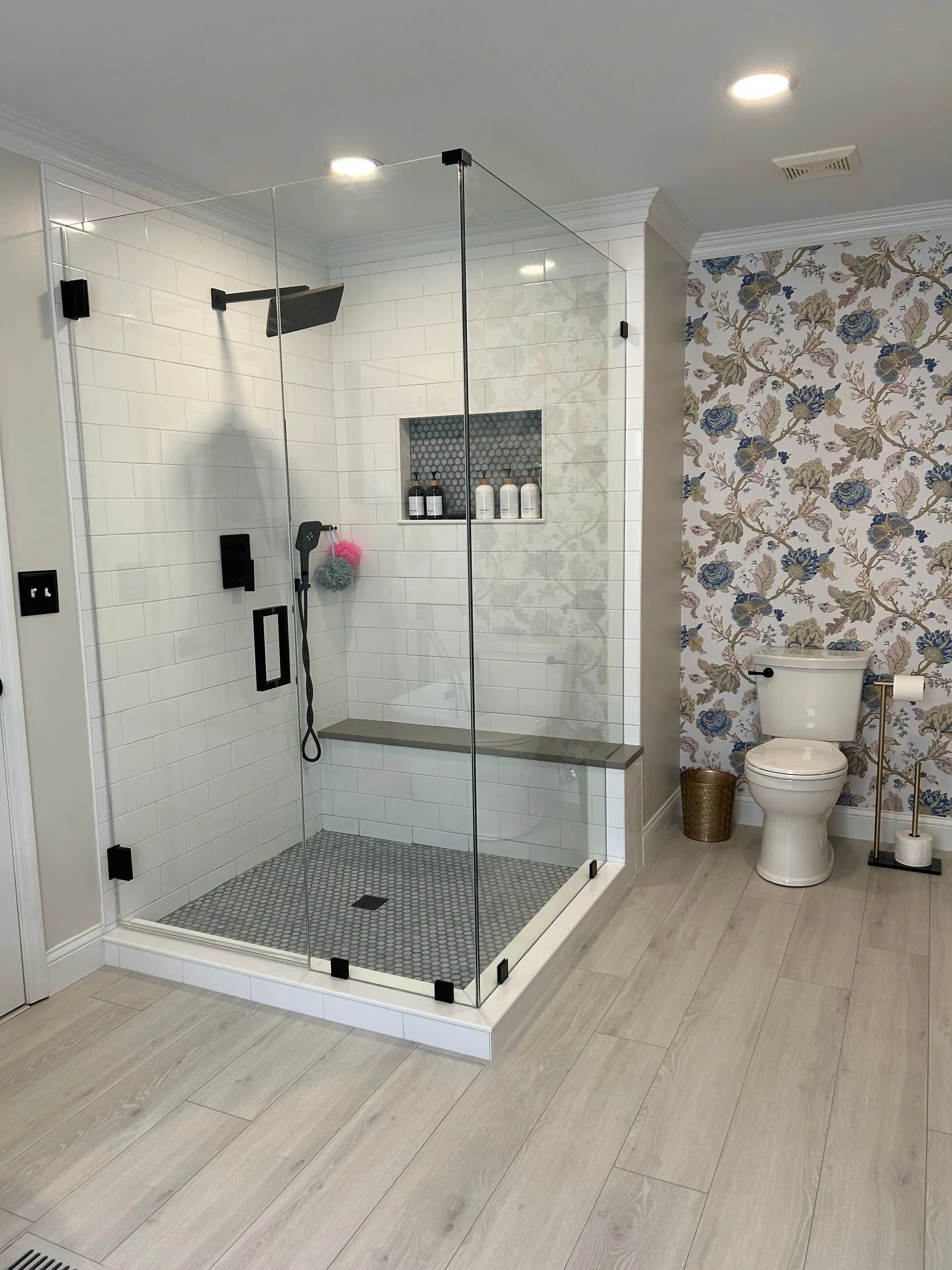 A modern bathroom with a glass-enclosed shower featuring patterned tile, a built-in bench, and floral wallpaper behind a toilet.