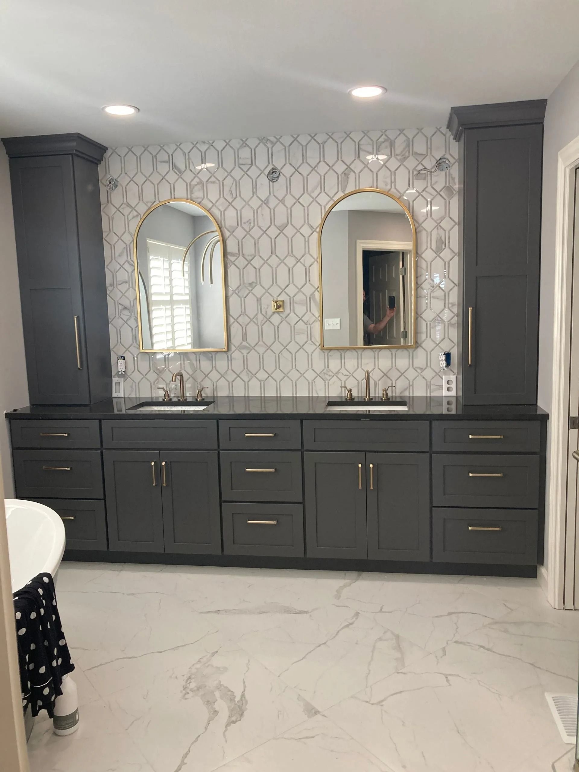Double vanity with dark gray cabinets, gold hardware, arched mirrors, and a white patterned tile backsplash in a bathroom.