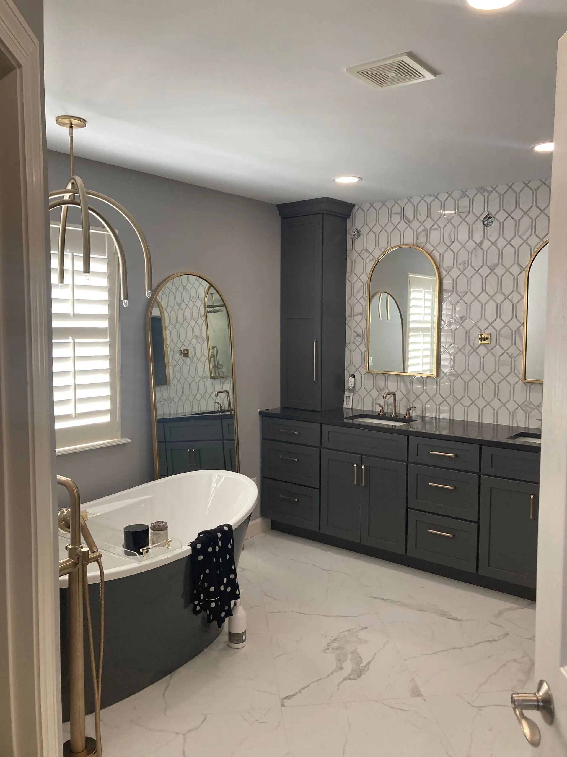 A modern bathroom featuring a charcoal vanity, a white freestanding tub, gold fixtures, and patterned wall tiles.