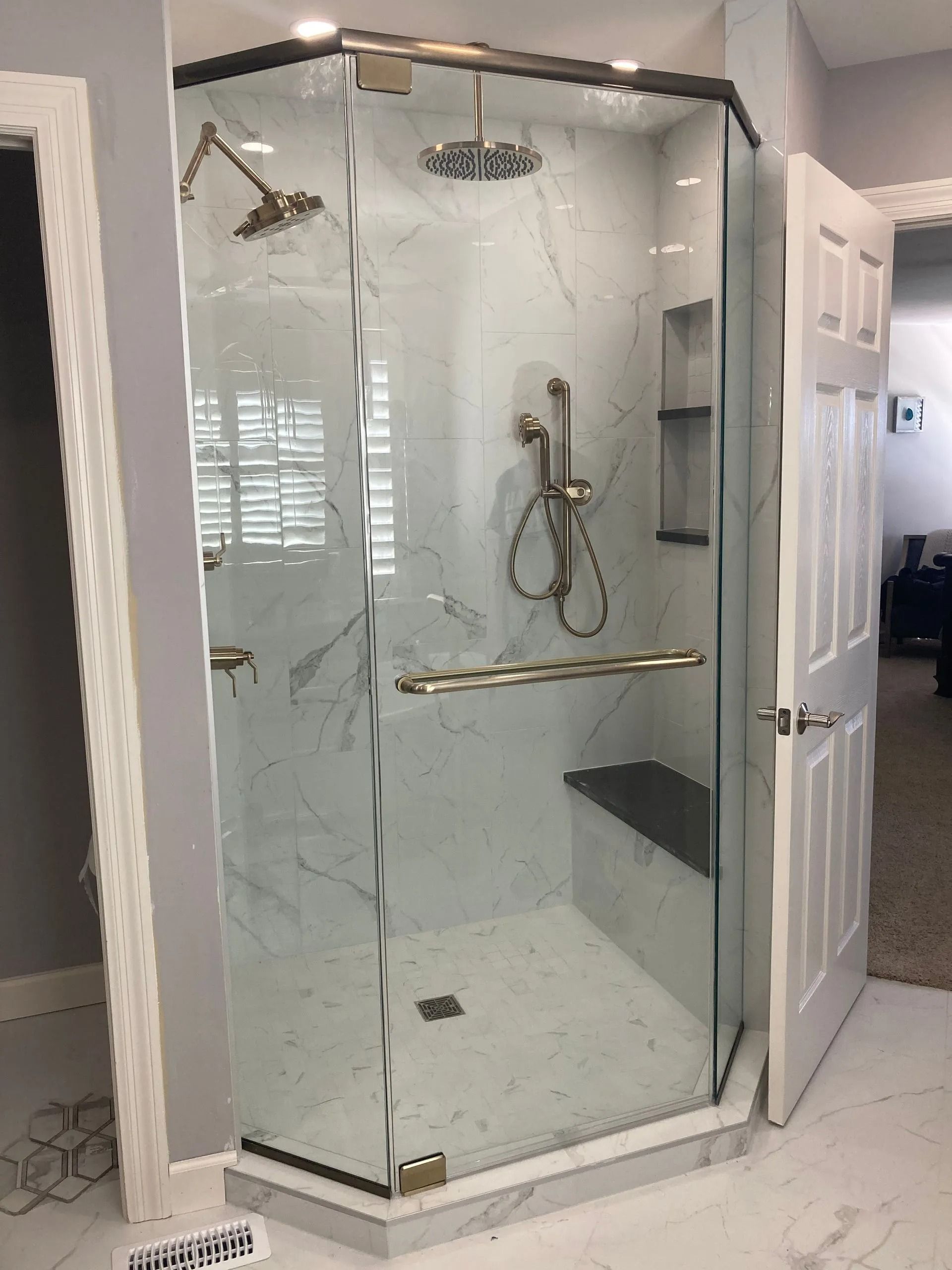 A neo-angle glass shower enclosure with white marble-look tiles, gold fixtures, a rainfall head, and a built-in bench.