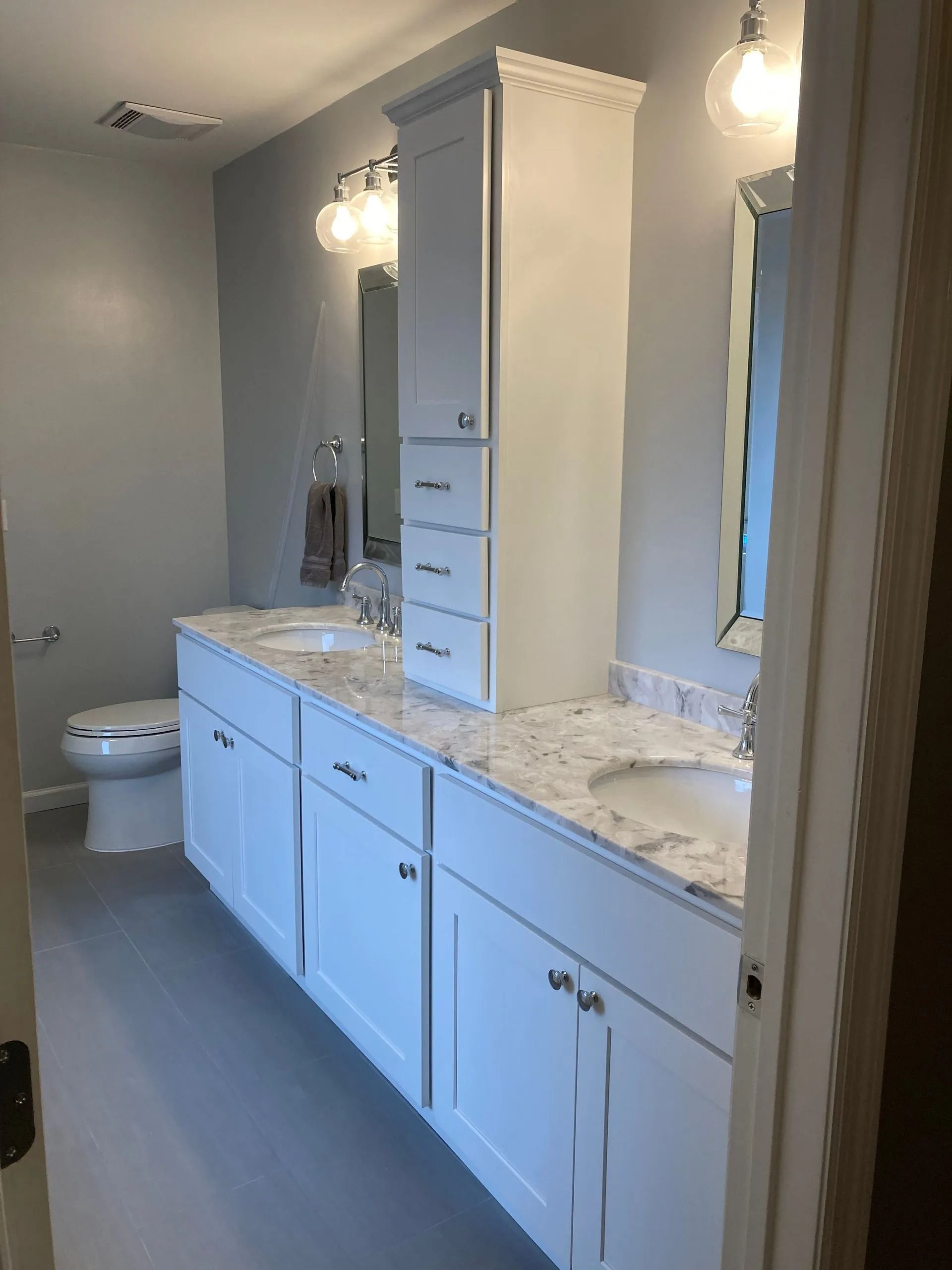 A modern bathroom featuring a white double vanity with a central tower cabinet, marble-look countertop, and gray walls.