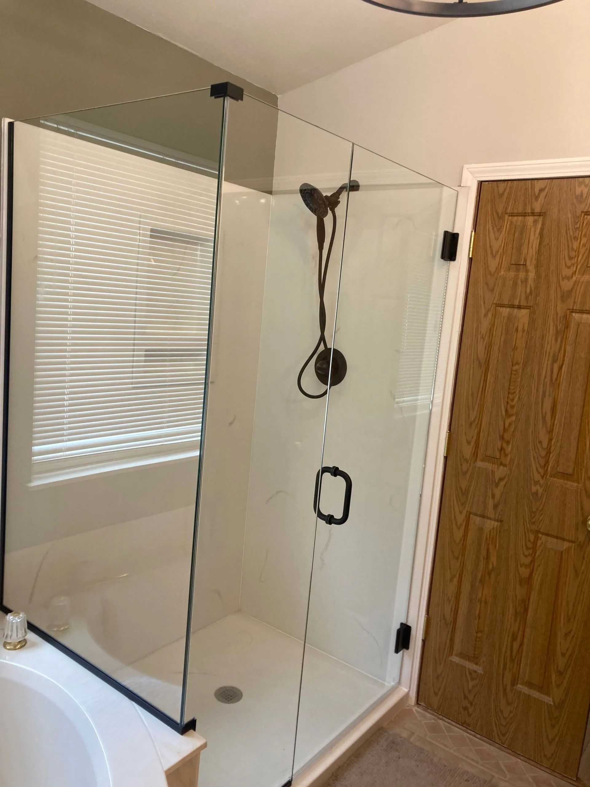 A glass shower enclosure with black hardware, centered in a bathroom next to a window with blinds and a wood door.