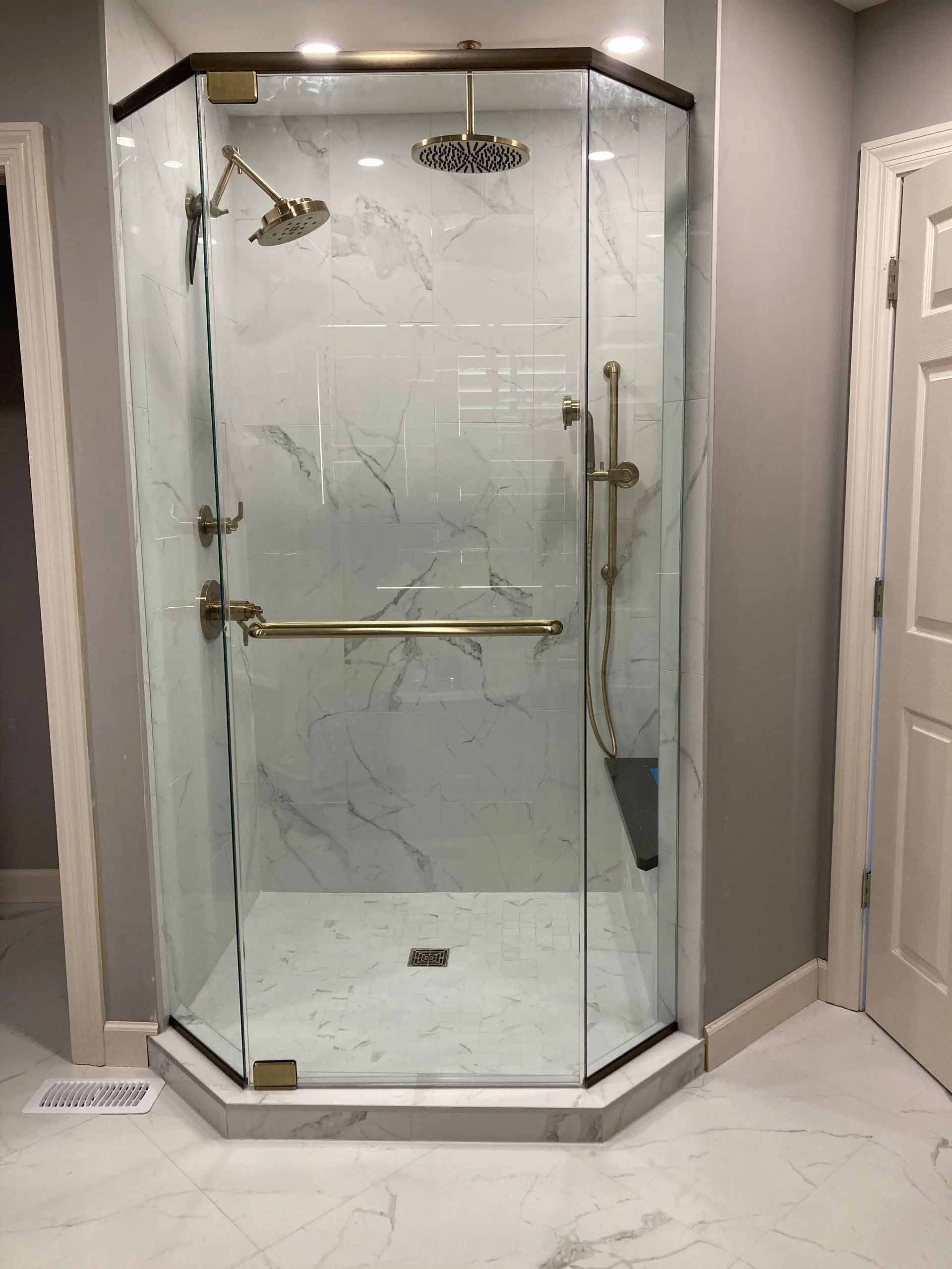 A modern walk-in shower with gold fixtures, glass panels, a rainfall showerhead, and a built-in seat against marble tiles.