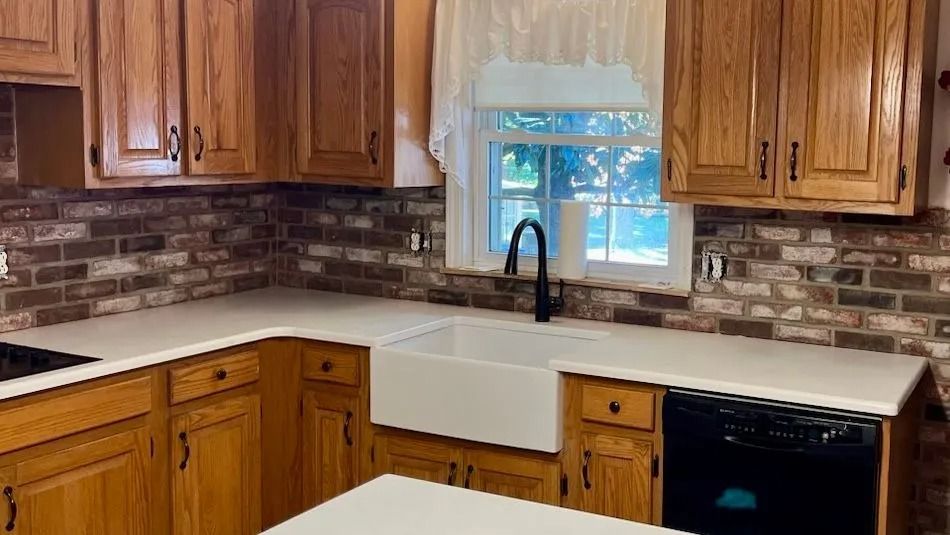 A kitchen with oak cabinets, brick backsplash, white farmhouse sink, and light countertops with a black dishwasher.