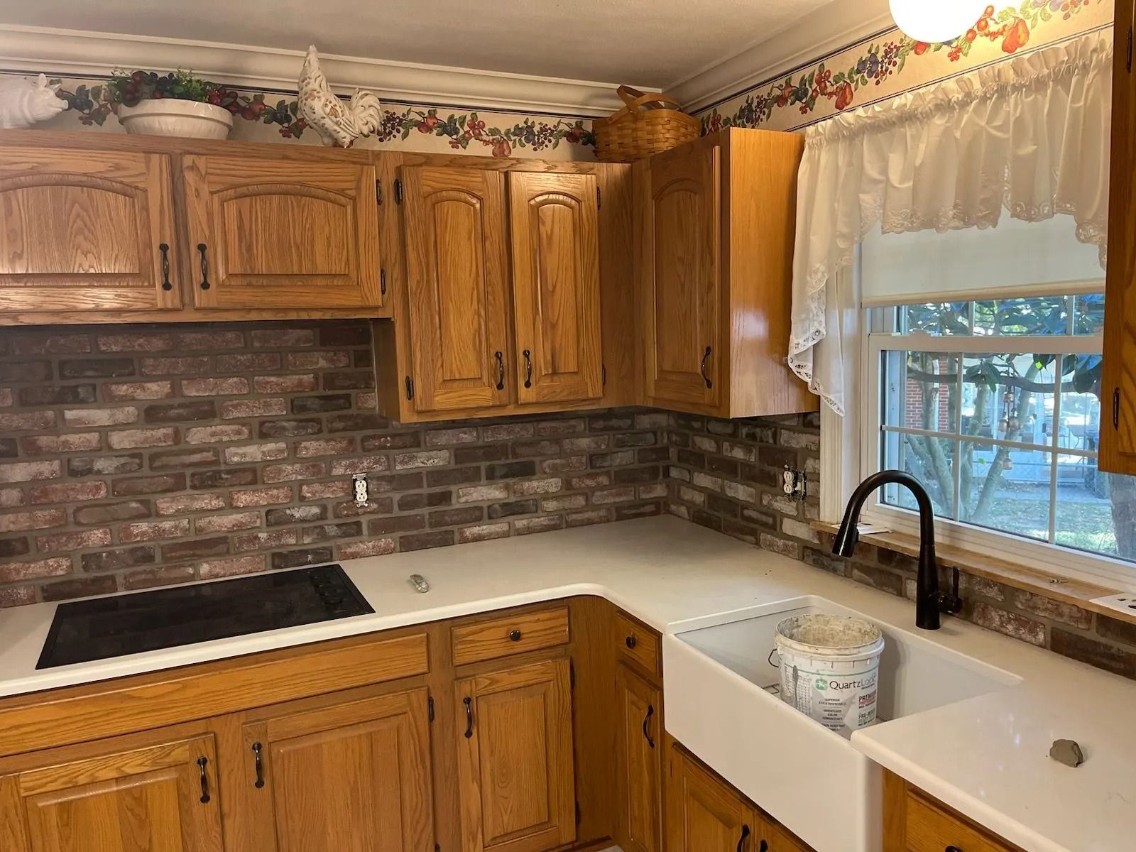 Kitchen with oak cabinets, a brick backsplash, white farmhouse sink, black faucet, and patterned border wallpaper.