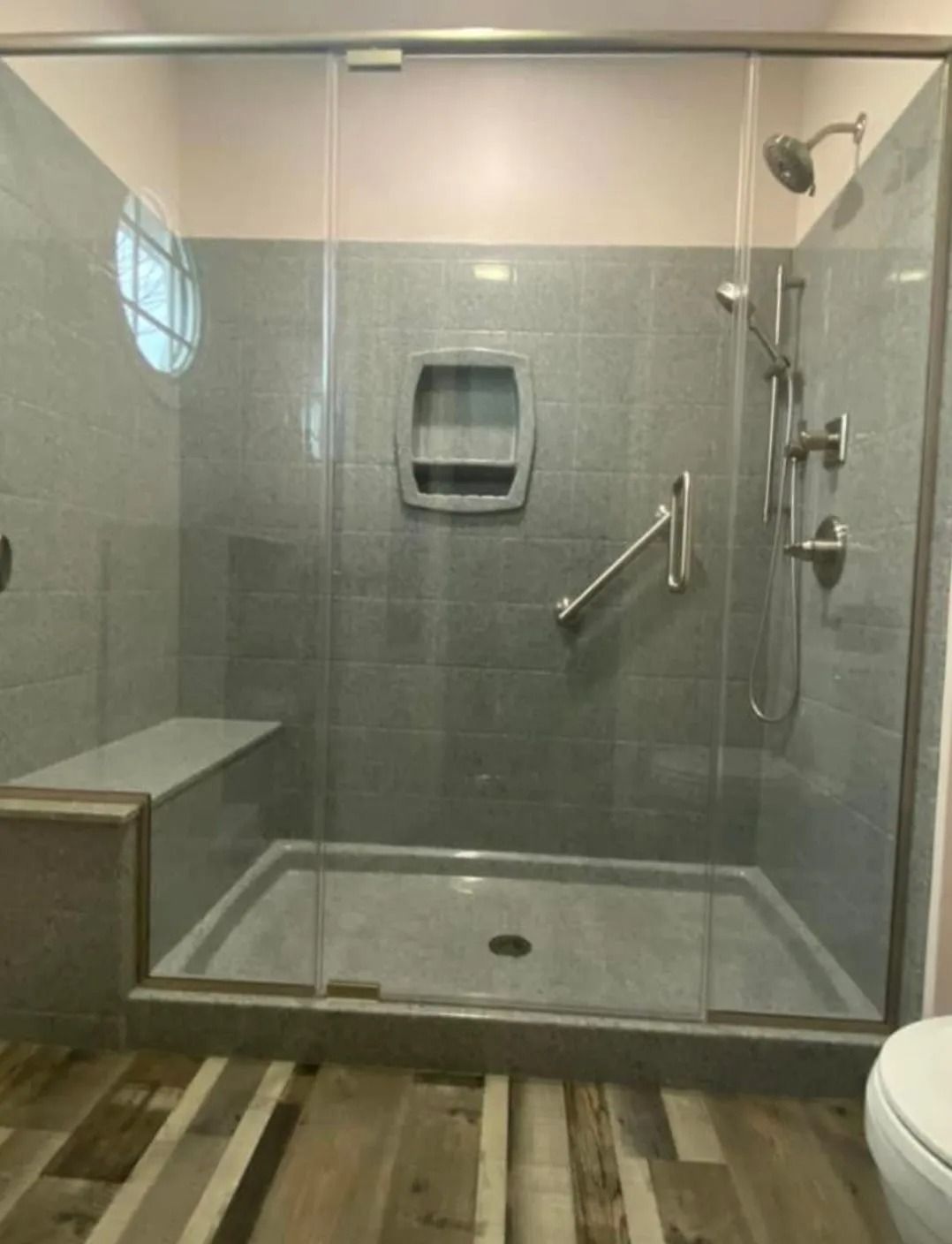 A glass-enclosed walk-in shower with grey tiled walls, a built-in bench, a silver shower head, and a metal grab bar.