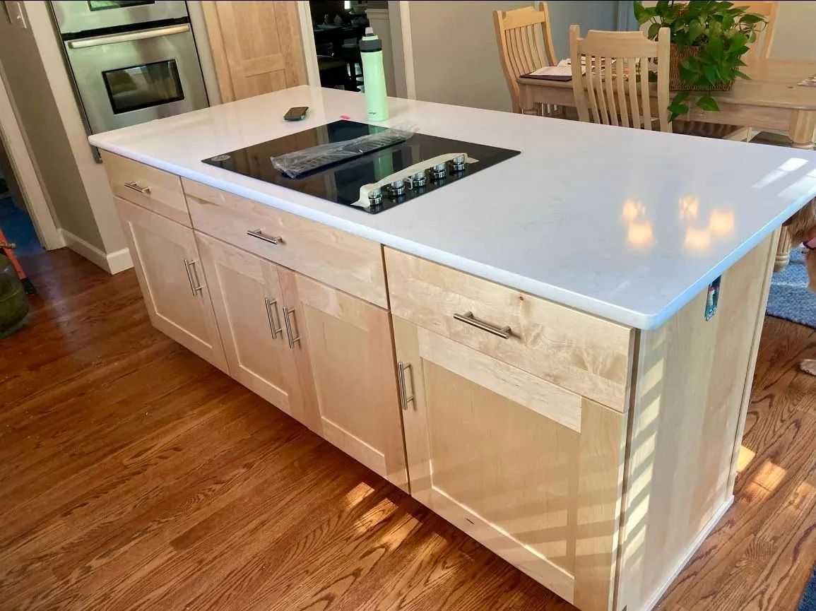 A light wood kitchen island with a white countertop and built-in stovetop, set on a hardwood floor in a home kitchen.