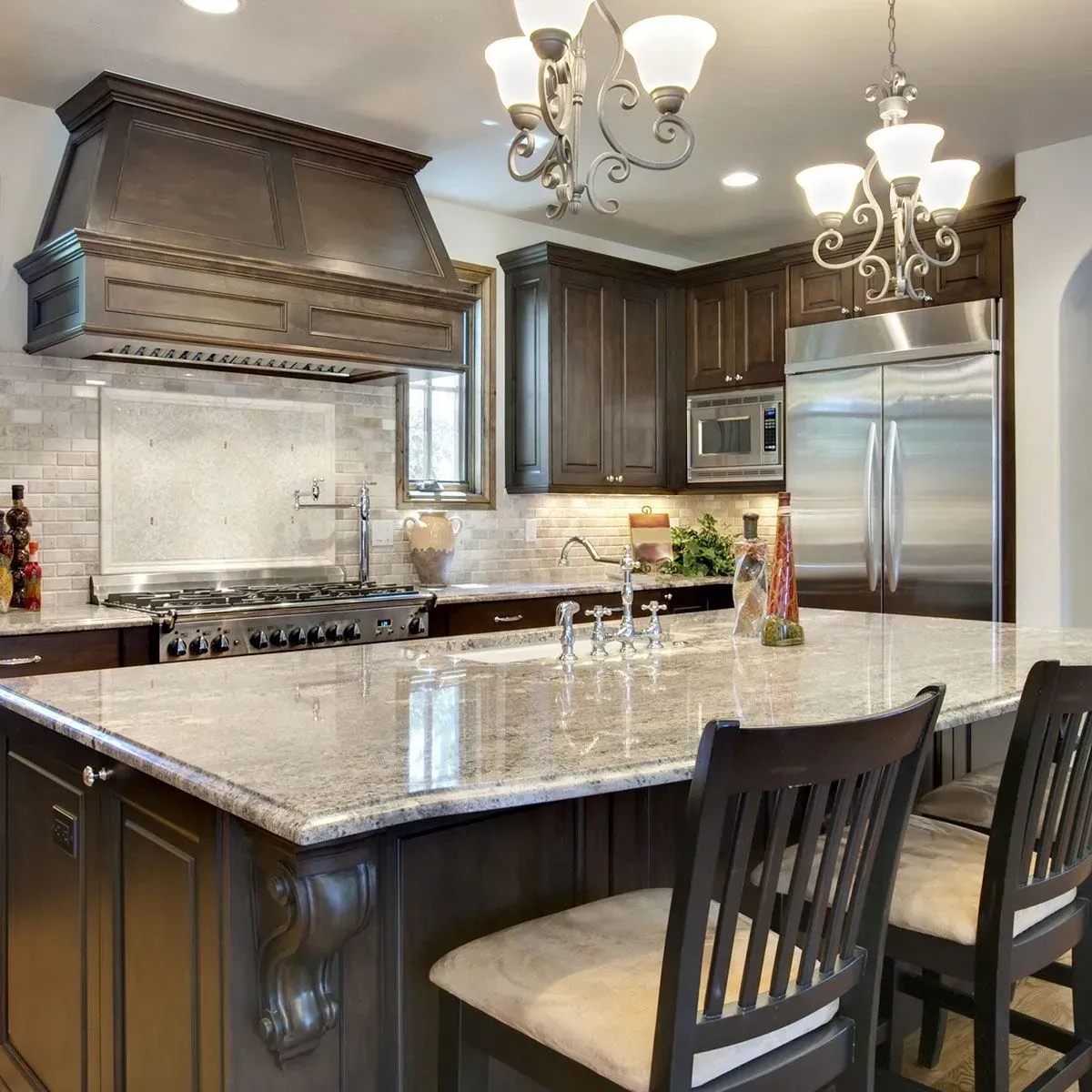 A spacious kitchen with dark wood cabinets, a large granite island, light brick backsplash, and two hanging chandeliers.