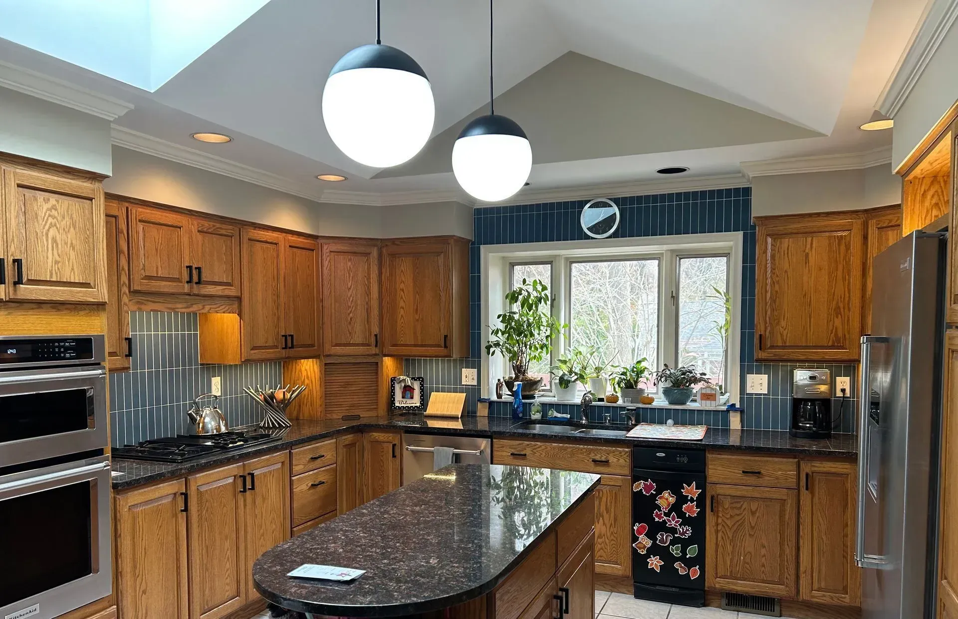 A kitchen with oak cabinets, a dark island, stainless steel appliances, and two globe lights beneath a vaulted ceiling.