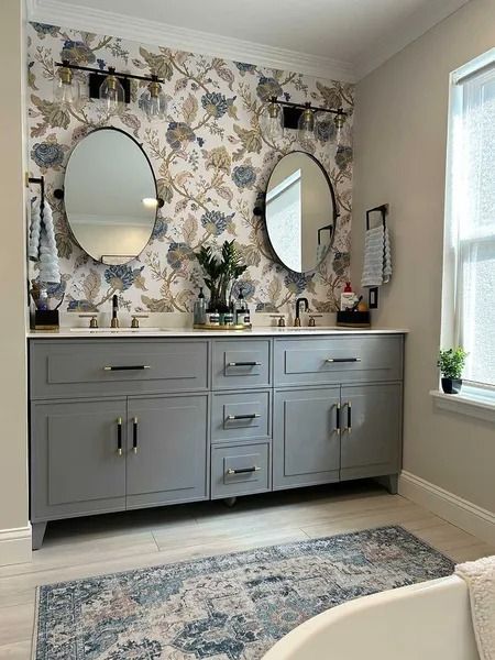 A bathroom vanity with grey cabinets, two oval mirrors, and floral wallpaper, set against a light-colored wall with a rug.
