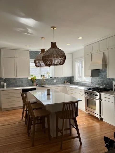 A white kitchen with a center island, wooden stools, woven pendant lights, and a stainless steel stove.
