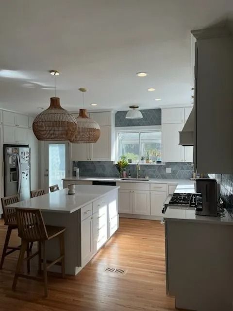 A white kitchen with a center island, woven pendant lights, stainless appliances, and light wood flooring.