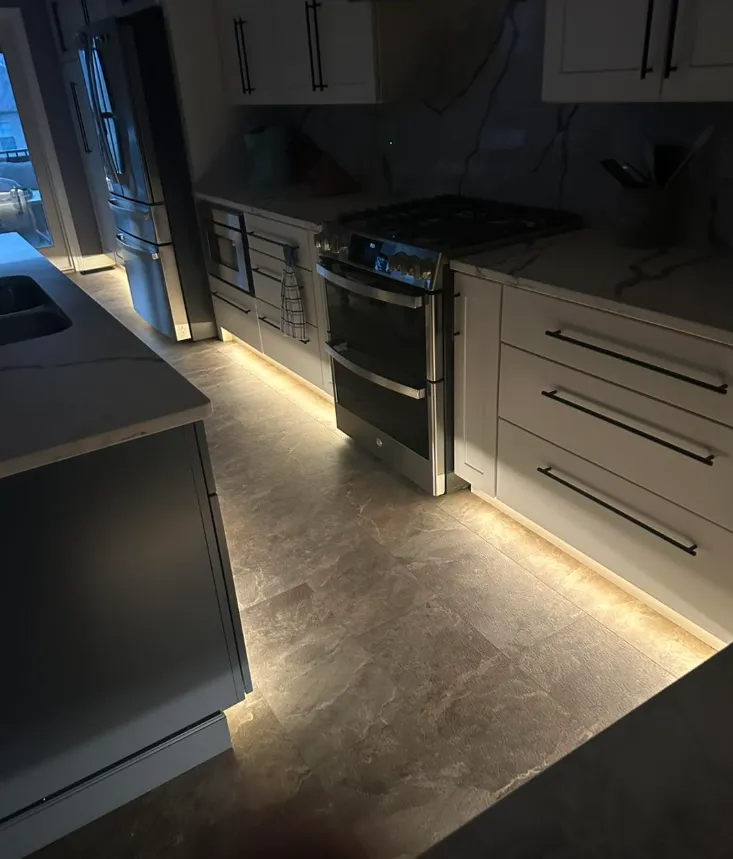 A modern kitchen features white cabinetry with under-cabinet and floor-level LED strip lighting illuminating the floor.