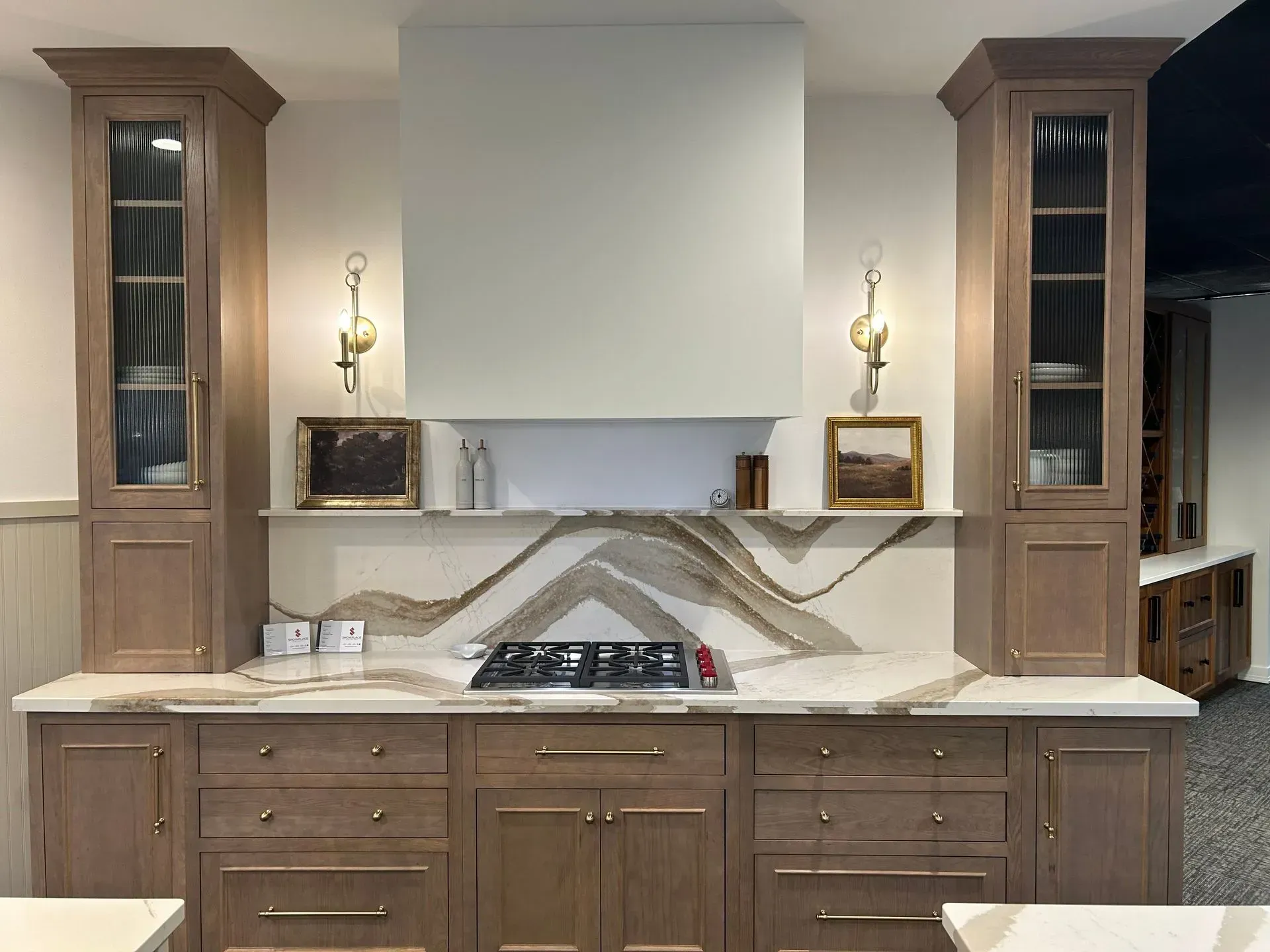 A kitchen range area with light wood cabinets, a white hood, stone backsplash, brass hardware, and wall sconces.