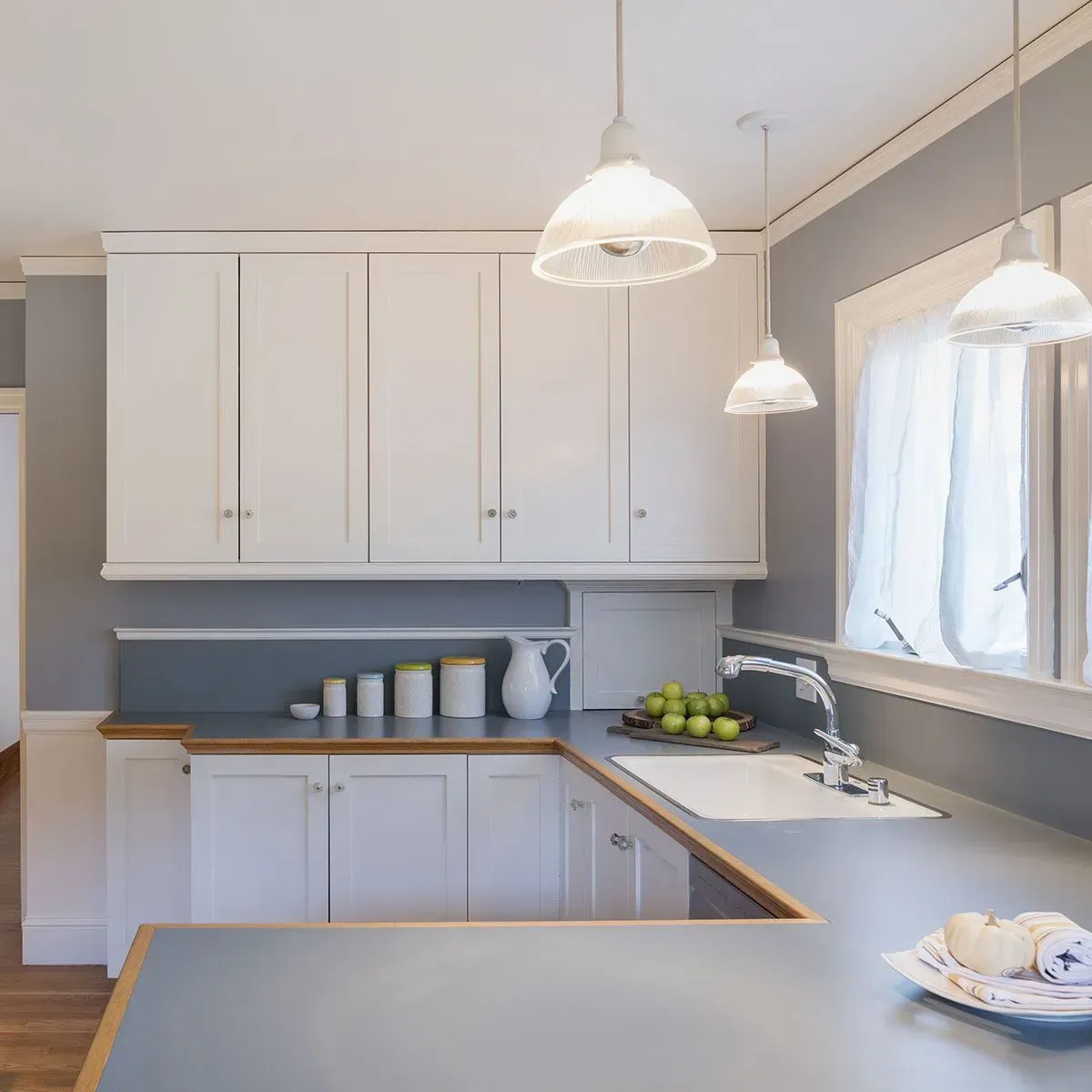 A bright kitchen featuring white cabinets, grey countertops, walls painted in a soft grey, and three glass pendant lights.
