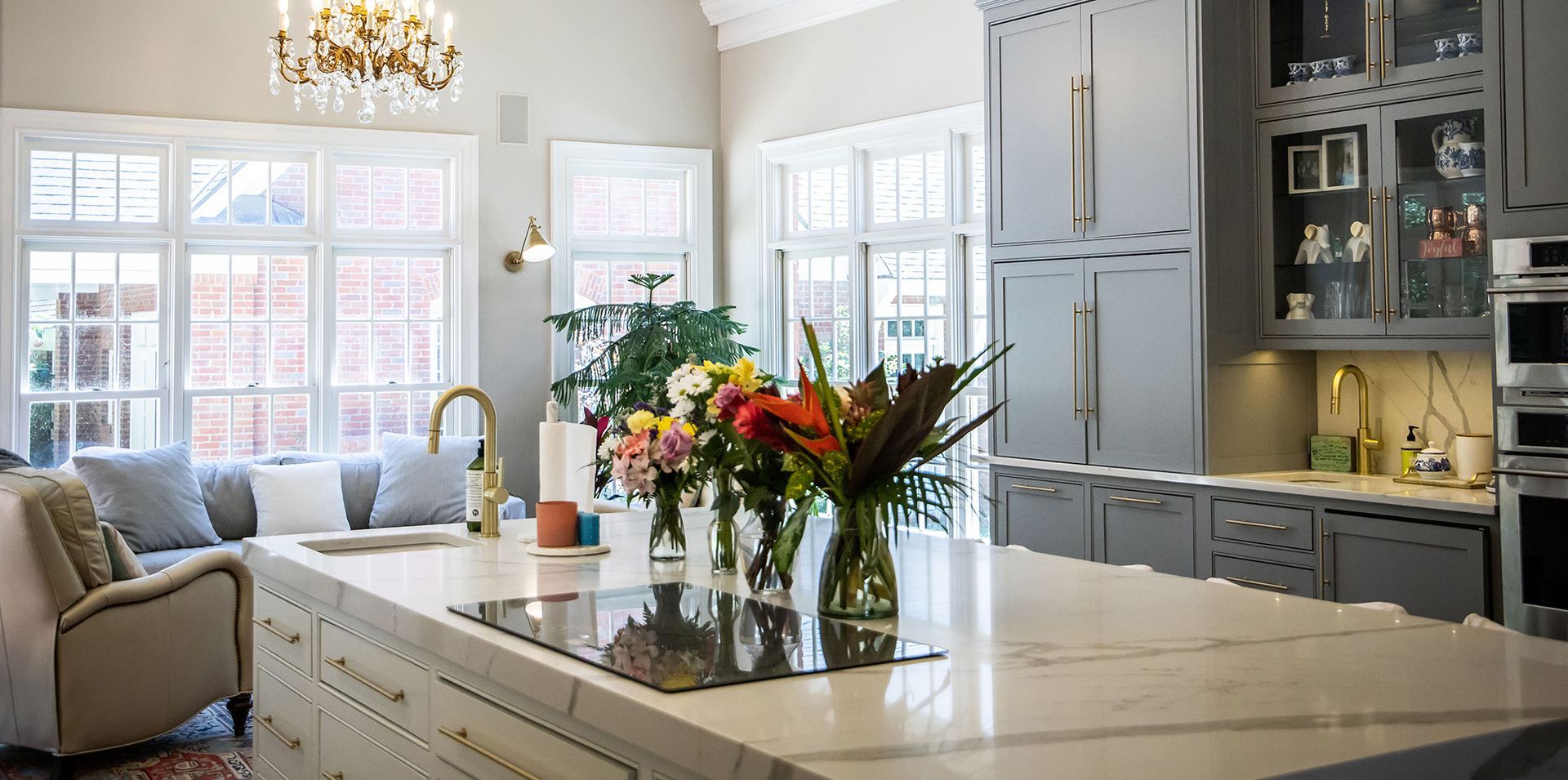 A bright, modern kitchen with a large marble island, floral centerpieces, gray cabinetry, and a cozy seating area.