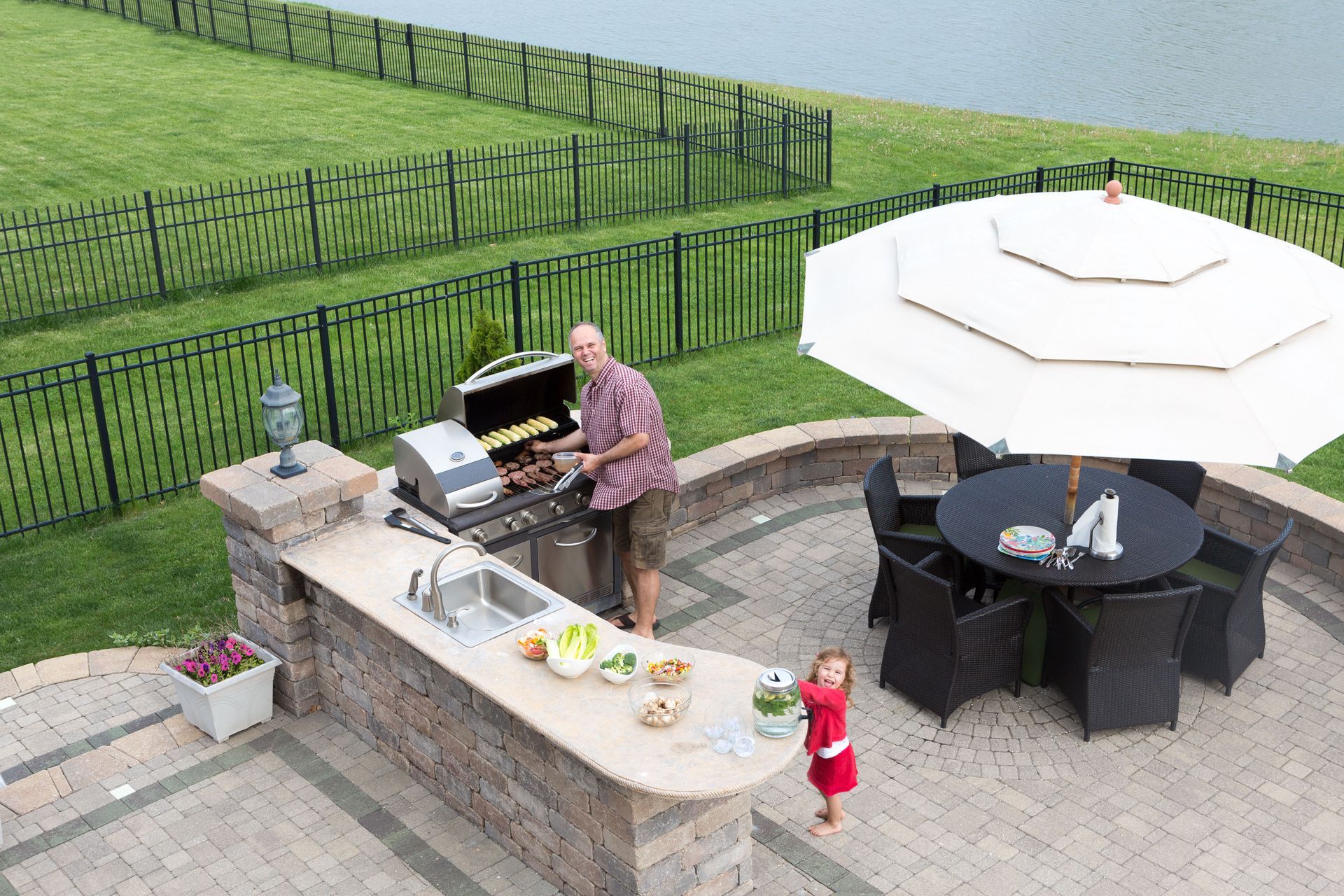 A person grills food at an outdoor kitchen while a child stands nearby on a stone patio with dining furniture.